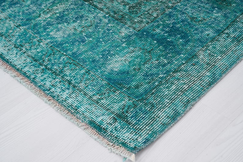 May include: Close-up of a teal blue rug with a faded, distressed pattern. The rug has a white fringe border.