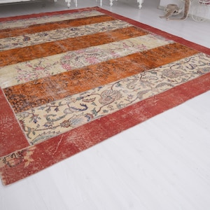 Puede incluir: Una alfombra rectangular con bandas horizontales alternas en color naranja quemado y crema. Las secciones crema muestran intrincados patrones florales en tonos azul, rosa y verde. Un borde ancho de color rojo s&oacute;lido enmarca todo el dise&ntilde;o.