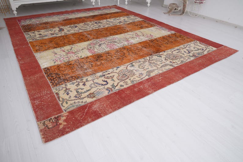 Puede incluir: Una alfombra rectangular con bandas horizontales alternas en color naranja quemado y crema. Las secciones crema muestran intrincados patrones florales en tonos azul, rosa y verde. Un borde ancho de color rojo s&oacute;lido enmarca todo el dise&ntilde;o.