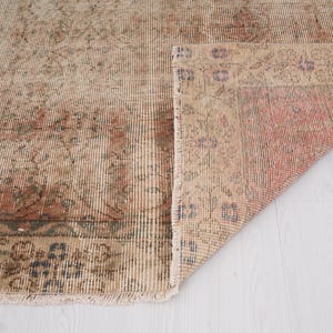 May include: A vintage-style rug with a distressed appearance. The rug features a beige and reddish-brown color palette with intricate floral patterns and a fringed edge. The corner is folded up, revealing the underside.