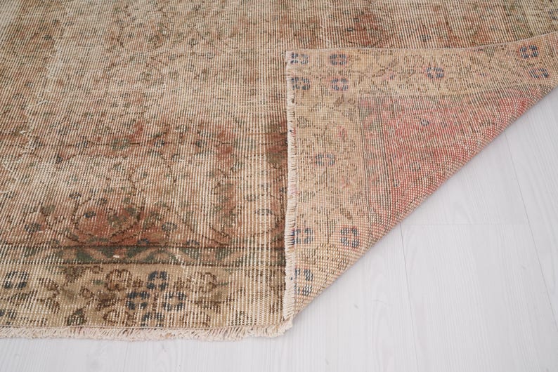 May include: A vintage-style rug with a distressed appearance. The rug features a beige and reddish-brown color palette with intricate floral patterns and a fringed edge. The corner is folded up, revealing the underside.