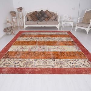 Puede incluir: Una alfombra rectangular grande con un borde rojo ancho y bandas horizontales alternas en color naranja quemado y crema. Las secciones crema est&aacute;n decoradas con intrincados patrones florales y de cachemira en tonos azul, marr&oacute;n y rosa.