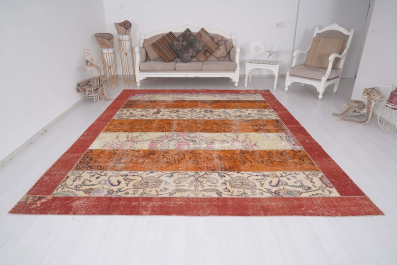 Puede incluir: Una alfombra rectangular grande con un borde rojo ancho y bandas horizontales alternas en color naranja quemado y crema. Las secciones crema est&aacute;n decoradas con intrincados patrones florales y de cachemira en tonos azul, marr&oacute;n y rosa.