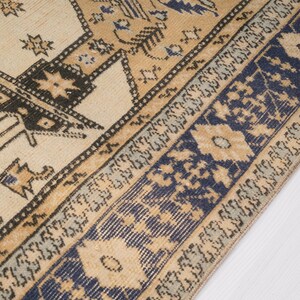May include: A vintage rug with a cream background and a blue and brown geometric pattern. The rug has a fringe border.