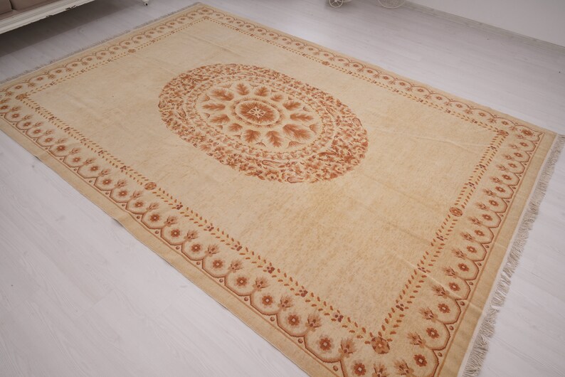 Puede incluir: Una alfombra beige con un patr&oacute;n floral marr&oacute;n. La alfombra tiene un borde de flecos y un gran medall&oacute;n ovalado en el centro.