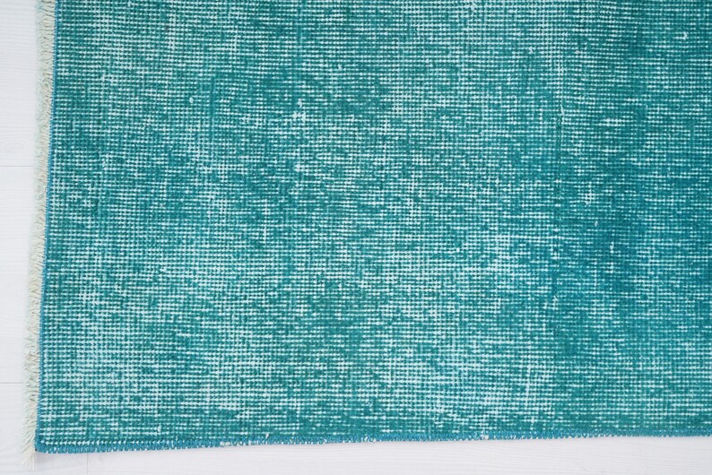 May include: A close-up of a teal blue rug with a woven texture. The rug has a white fringe along the edges.