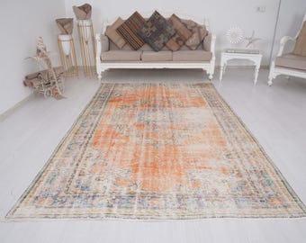 May include: A rectangular area rug featuring a prominent orange central field with faded blue and cream floral patterns. The rug has a detailed border with geometric and botanical motifs in muted tones, finished with short fringe on the ends.