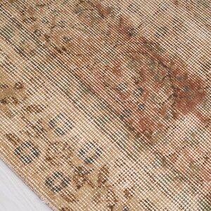 May include: Close-up of a distressed, vintage rug with a beige and brown color palette. The rug features faded floral patterns and a textured surface, showcasing a worn, antique aesthetic. The design includes accents of red and green.