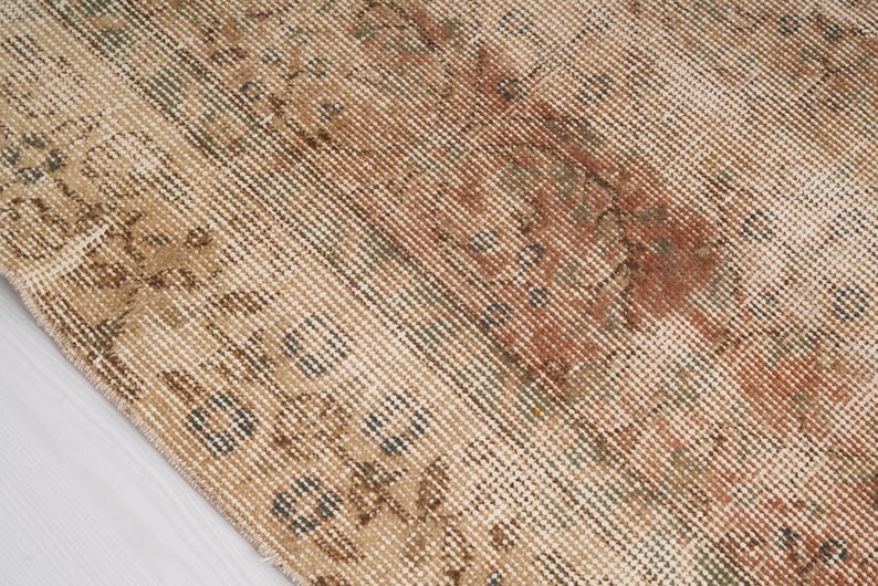 May include: Close-up of a distressed, vintage rug with a beige and brown color palette. The rug features faded floral patterns and a textured surface, showcasing a worn, antique aesthetic. The design includes accents of red and green.