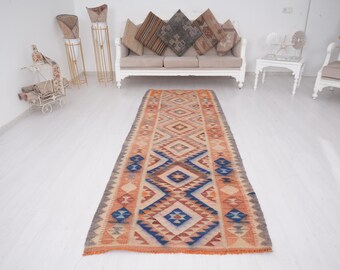 May include: A long, rectangular rug with a geometric pattern in shades of orange, blue, and beige. The rug has a fringe border and is made of wool.