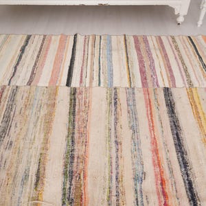 May include: A rectangular area rug featuring a woven striped pattern with a variety of colors including cream, beige, red, blue, yellow, and black. The rug is composed of multiple vertical bands of varying widths and textures throughout the surface.