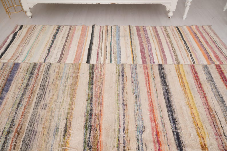 May include: A rectangular area rug featuring a woven striped pattern with a variety of colors including cream, beige, red, blue, yellow, and black. The rug is composed of multiple vertical bands of varying widths and textures throughout the surface.