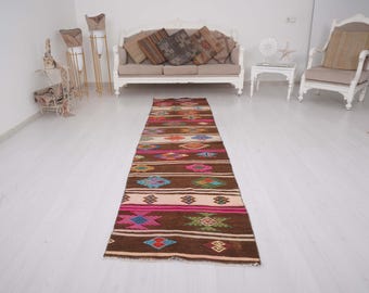 May include: A long, rectangular runner rug featuring a dark brown base with horizontal stripes in cream and bright pink. The rug is decorated with a repeating pattern of geometric diamond shapes in vibrant colors including blue, green, and orange.