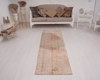 May include: A long, rectangular, off-white runner rug with a faded, distressed, and worn appearance. The rug has a subtle, faded, peachy-pink color throughout. The rug is lying on a white wood floor.