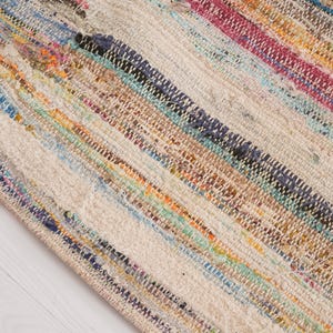 May include: A woven area rug featuring a multi-colored striped pattern with bands of pink, blue, orange, yellow, and dark green interspersed with a neutral off-white base. The textile displays a textured, hand-loomed appearance with visible fibers.