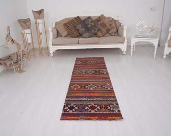 May include: A rectangular woven runner rug featuring a repeating pattern of geometric shapes and horizontal stripes in shades of orange, red, purple, green, and dark blue. The rug has a flat-weave texture with a multi-colored design throughout.