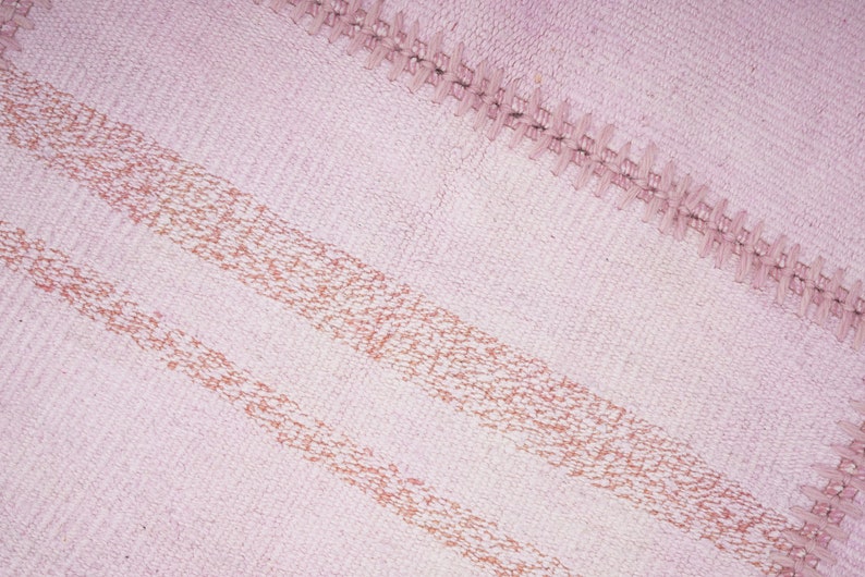 May include: A pink woven rug with two horizontal stripes of a darker pink color. The rug has a fringe border.