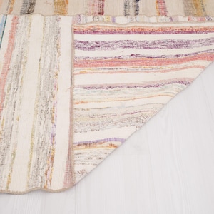 May include: A woven area rug featuring a multi-colored striped pattern with shades of purple, pink, yellow, blue, and cream. The rug is folded at one corner, revealing the underside which displays a similar striped design in lighter tones.