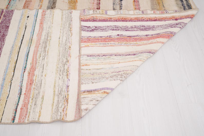May include: A woven area rug featuring a multi-colored striped pattern with shades of purple, pink, yellow, blue, and cream. The rug is folded at one corner, revealing the underside which displays a similar striped design in lighter tones.