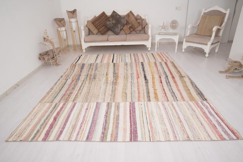 May include: A rectangular area rug featuring a multi-colored striped pattern with a palette of cream, beige, pink, yellow, and dark brown tones. The rug displays a woven texture with vertical lines running across the entire surface of the piece.