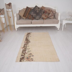 May include: A rectangular beige rug with a decorative border featuring floral and geometric patterns in shades of brown, blue, and orange. The rug is placed on a light-colored wooden floor, with a sofa and decorative items in the background.