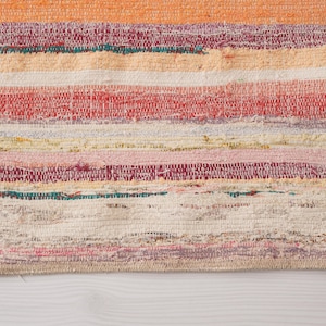 May include: A rectangular woven rug featuring horizontal stripes in various colors including orange, red, pink, yellow, white, and teal. The fabric has a textured, multi-colored appearance with visible woven threads and a finished edge border.