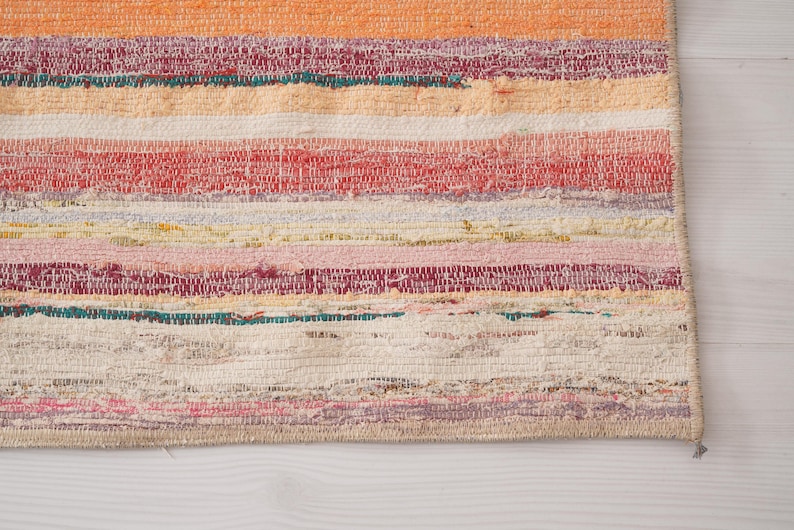 May include: A rectangular woven rug featuring horizontal stripes in various colors including orange, red, pink, yellow, white, and teal. The fabric has a textured, multi-colored appearance with visible woven threads and a finished edge border.