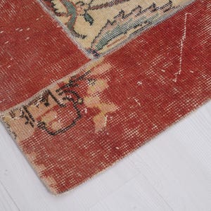 Puede incluir: Una alfombra tejida que presenta un borde rojo intenso prominente con una textura desgastada y descolorida. La secci&oacute;n central muestra un campo de color crema adornado con intrincados patrones florales en tonos apagados de azul, verde y bronceado, mostrando signos de desgaste.