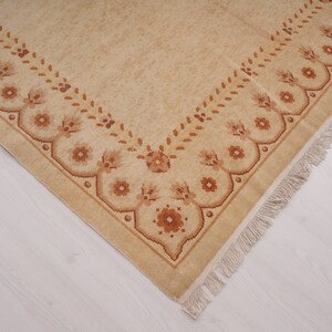 Puede incluir: Una alfombra beige con un borde de patr&oacute;n floral marr&oacute;n y flecos.