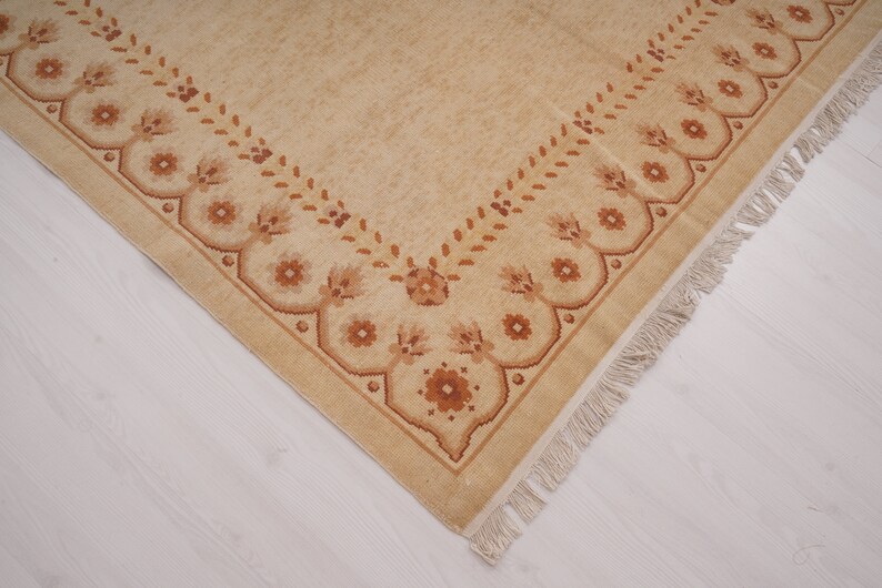Puede incluir: Una alfombra beige con un borde de patr&oacute;n floral marr&oacute;n y flecos.