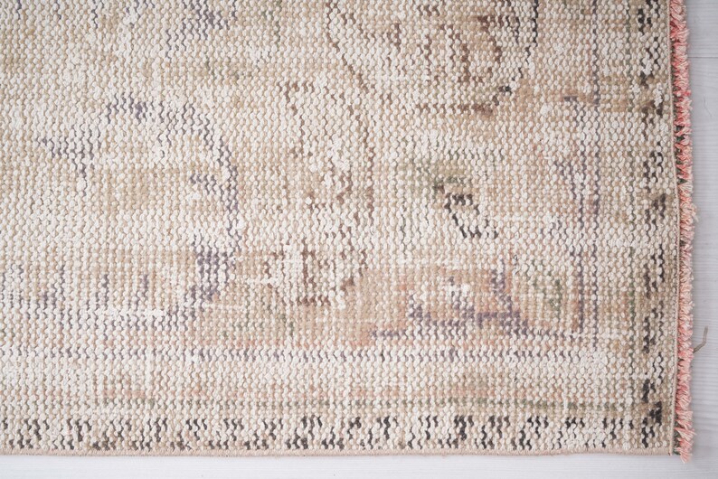 Op de afbeelding: Een close-up van een beige kleed met een subtiel patroon. Het kleed heeft een roze franje langs de rand.