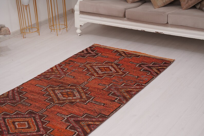 May include: A long, narrow rug with a geometric pattern in shades of orange, brown, and white. The rug has a fringed edge and is made of a soft, woven material.