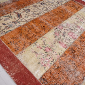 Puede incluir: Una alfombra rectangular con un dise&ntilde;o de patchwork de franjas verticales alternas. El patr&oacute;n consiste en secciones de color naranja quemado s&oacute;lido y paneles de color crema decorados con intrincados motivos florales en tonos rosa, azul y verde.