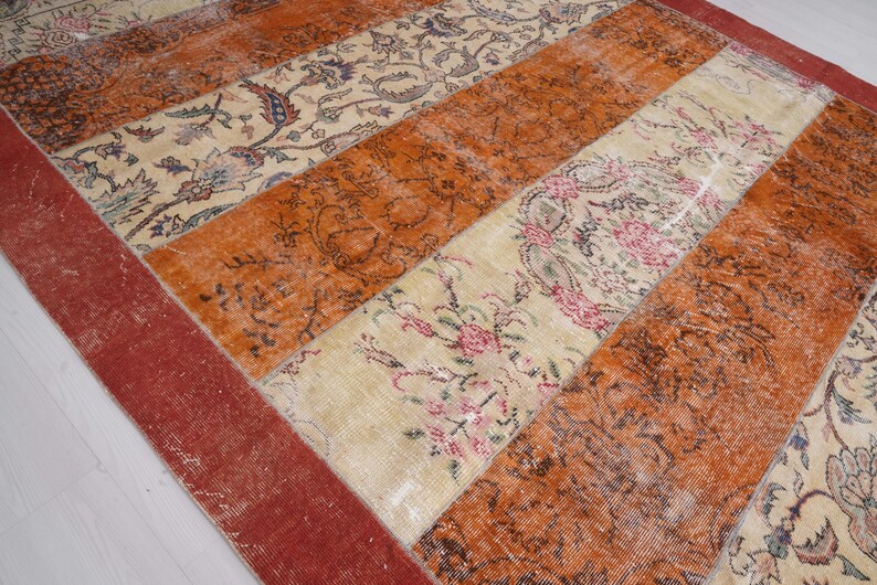Puede incluir: Una alfombra rectangular con un dise&ntilde;o de patchwork de franjas verticales alternas. El patr&oacute;n consiste en secciones de color naranja quemado s&oacute;lido y paneles de color crema decorados con intrincados motivos florales en tonos rosa, azul y verde.
