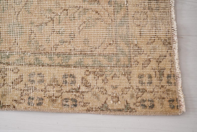 May include: Close-up of a section of a beige and green patterned rug. The rug has a distressed, vintage look with intricate floral and geometric designs. The edges are frayed.