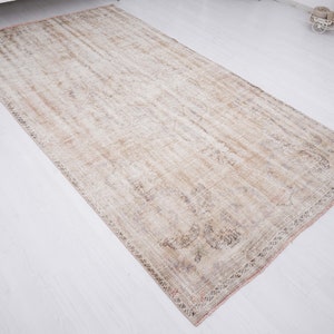 Op de afbeelding: Een vintage tapijt in een verbleekte beige en bruine kleurstelling. Het tapijt heeft een verweerde uitstraling met een subtiel patroon over de gehele oppervlakte. Het tapijt heeft een gefranst randje.