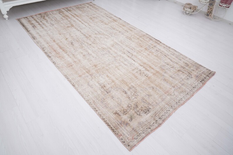 Op de afbeelding: Een vintage tapijt in een verbleekte beige en bruine kleurstelling. Het tapijt heeft een verweerde uitstraling met een subtiel patroon over de gehele oppervlakte. Het tapijt heeft een gefranst randje.