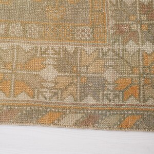 May include: Close-up of a section of a vintage rug with a geometric pattern. The rug features a blend of muted colors, including olive green, beige, and touches of orange and brown. The design includes repeating floral and geometric shapes.