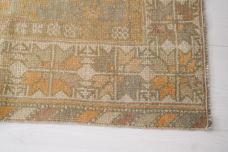 May include: Close-up of a section of a vintage rug with a geometric pattern. The rug features a blend of muted colors, including olive green, beige, and touches of orange and brown. The design includes repeating floral and geometric shapes.