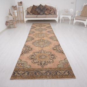 May include: A long, rectangular rug with an intricate, symmetrical pattern in shades of peach, brown, and olive green. The rug features a central medallion design and ornate borders, suitable for a hallway or living space.