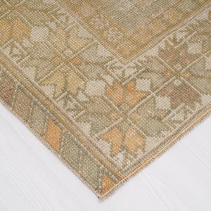 May include: Close-up of a corner of a vintage rug with a geometric pattern. The rug features a beige and olive green color palette with accents of orange and brown. The rug's design includes floral and geometric shapes.