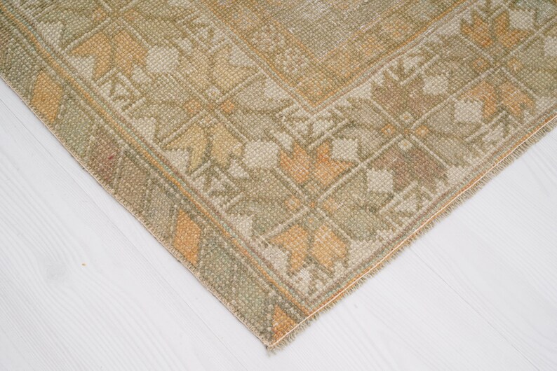 May include: Close-up of a corner of a vintage rug with a geometric pattern. The rug features a beige and olive green color palette with accents of orange and brown. The rug's design includes floral and geometric shapes.