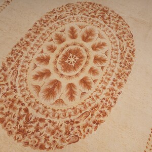 Puede incluir: Una alfombra beige con un gran dise&ntilde;o ovalado marr&oacute;n en el centro. El dise&ntilde;o ovalado presenta un patr&oacute;n repetitivo de hojas y espirales. La alfombra tiene un borde de dise&ntilde;os florales marrones m&aacute;s peque&ntilde;os.