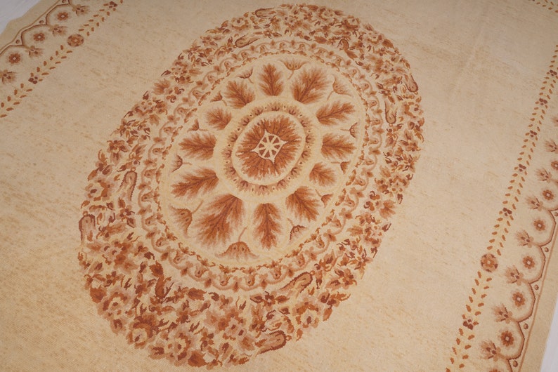 Puede incluir: Una alfombra beige con un gran dise&ntilde;o ovalado marr&oacute;n en el centro. El dise&ntilde;o ovalado presenta un patr&oacute;n repetitivo de hojas y espirales. La alfombra tiene un borde de dise&ntilde;os florales marrones m&aacute;s peque&ntilde;os.