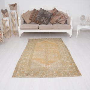 May include: An antique, rectangular rug with a central medallion design in shades of gold and beige. The rug has a distressed, vintage appearance, with a detailed border and a faded look. The rug is placed on a white floor.