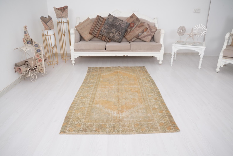 May include: An antique, rectangular rug with a central medallion design in shades of gold and beige. The rug has a distressed, vintage appearance, with a detailed border and a faded look. The rug is placed on a white floor.