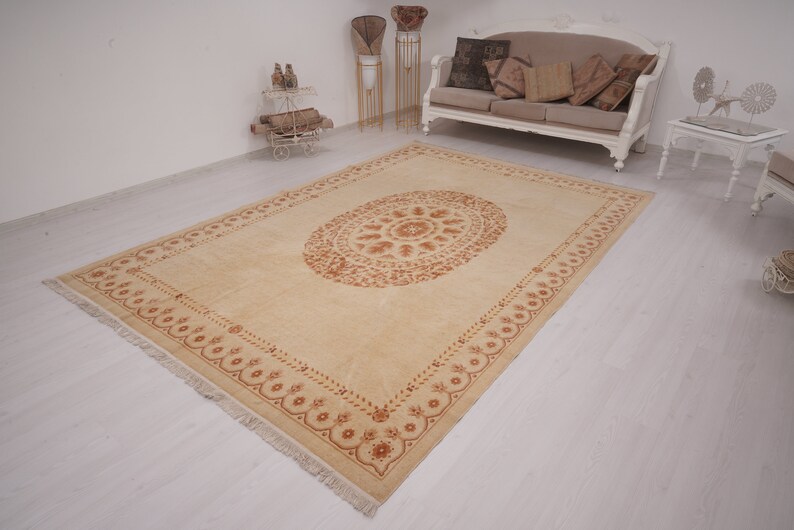 Puede incluir: Una alfombra beige con un gran dise&ntilde;o ovalado intrincado en un tono marr&oacute;n m&aacute;s oscuro. La alfombra tiene un borde con flecos.