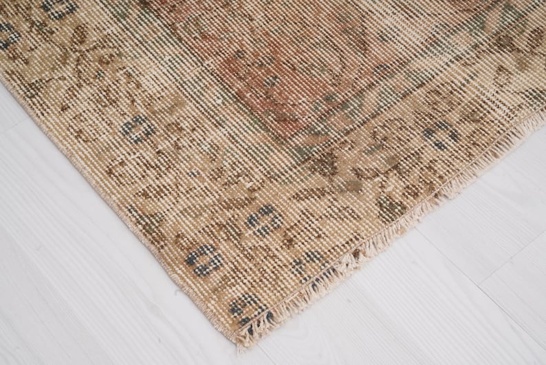 May include: Close-up of a distressed, vintage rug with a faded, textured appearance. The rug features a beige and brown color palette with hints of red and blue. The edges are frayed, adding to the antique aesthetic.