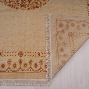 Puede incluir: Una alfombra beige con un patr&oacute;n floral marr&oacute;n. La alfombra tiene un borde de flecos y est&aacute; doblada por la mitad.