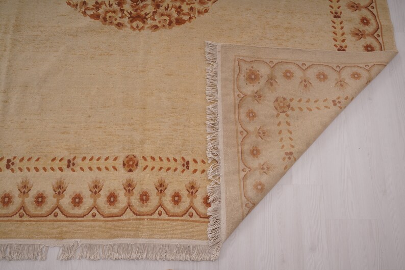 Puede incluir: Una alfombra beige con un patr&oacute;n floral marr&oacute;n. La alfombra tiene un borde de flecos y est&aacute; doblada por la mitad.